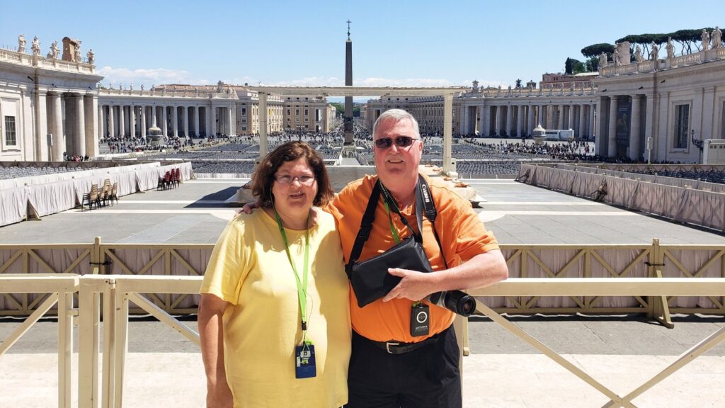 A smiling couple standing in St. Peter's Square in the Vatican with the Egyptian obelisk in the background.