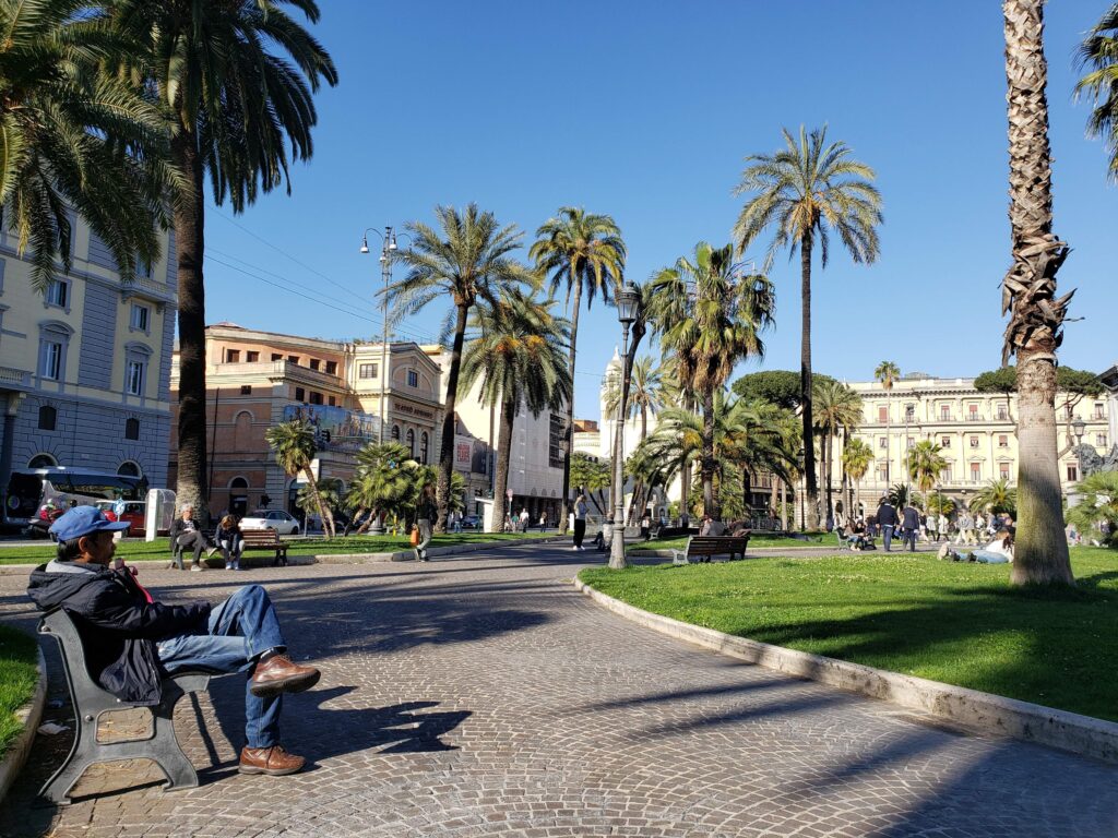 People relaxing on park benches in Piazza Cavour under tall palm trees.