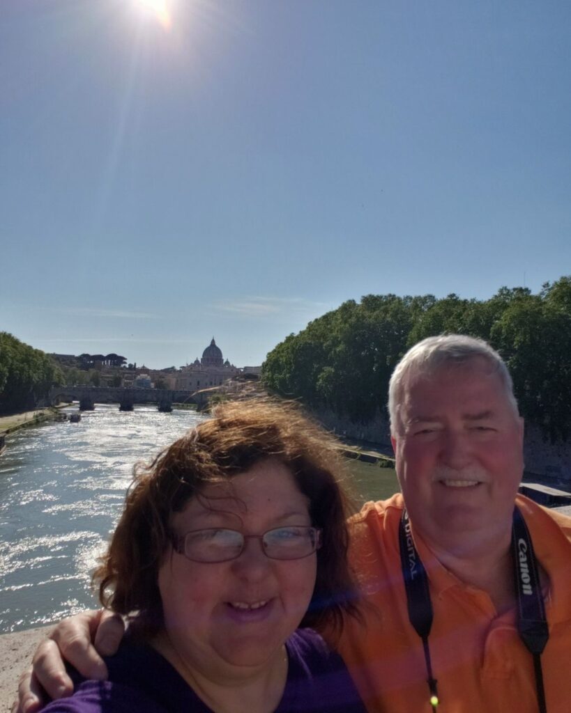 Tara and Mike taking a selfie on a bridge in Rome with the river and St. Peter's Basilica in the background.