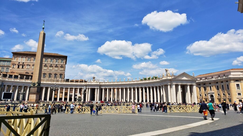 Wide view of St. Peter's Square in Vatican City featuring the central obelisk and colonnades under a bright blue sky.