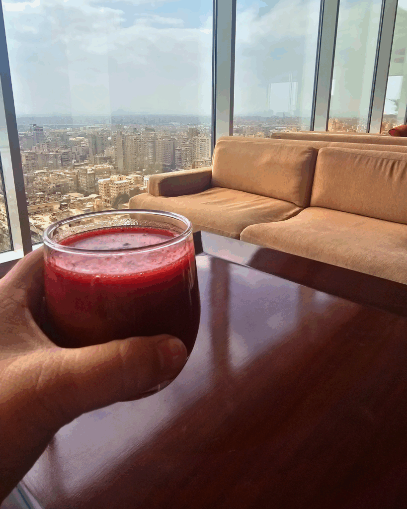 A glass of deep red Karkadeh held up against a panoramic view of Cairo from a high-rise hotel lounge.