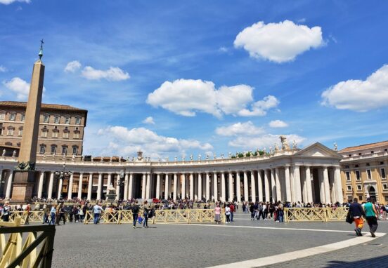 Wide view of St. Peter's Square in Vatican City featuring the central obelisk and colonnades under a bright blue sky.